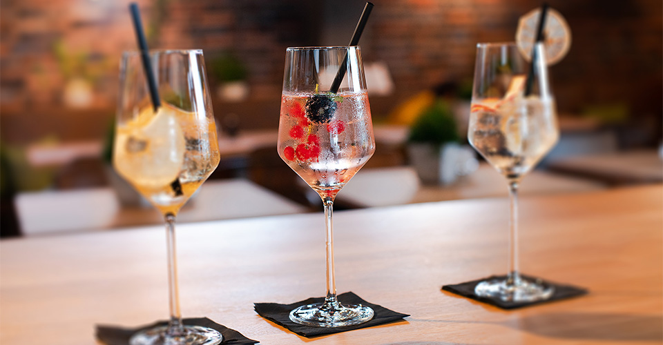 Drei Cocktails in eleganten Gläsern auf einer Holzoberfläche mit Früchten und Strohhalmen.