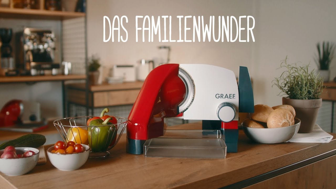 Küchenszene mit Aufschnittmaschine, Gemüse und Brötchen auf dem Tisch.