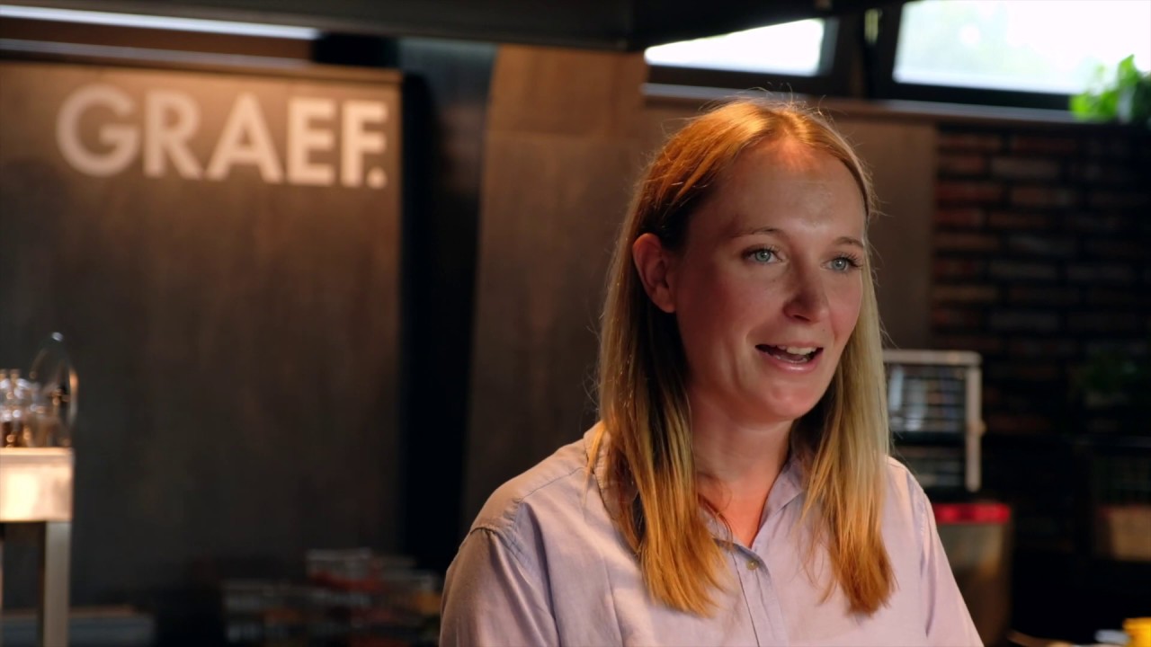 Frau spricht vor GRAEF-Logo, blondes Haar, blauäugig, Hemd, Innenraum, Tag