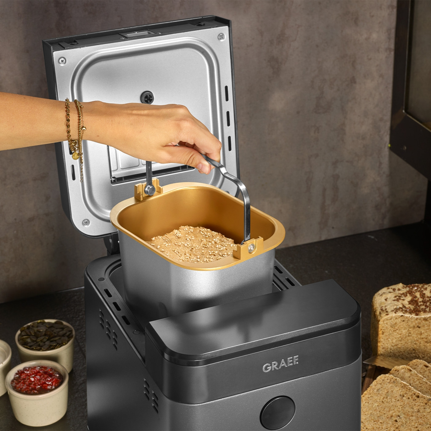 Bread maker with ingredients and hand holding the container, GRAEF logo visible