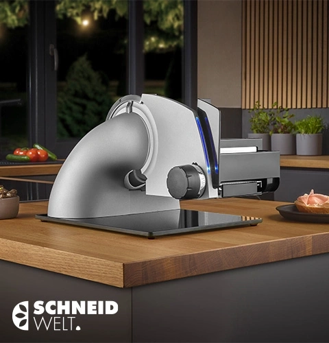 Schneidewelt Allesschneider auf einer Holzoberfläche mit Tomaten und Gurken im Hintergrund.