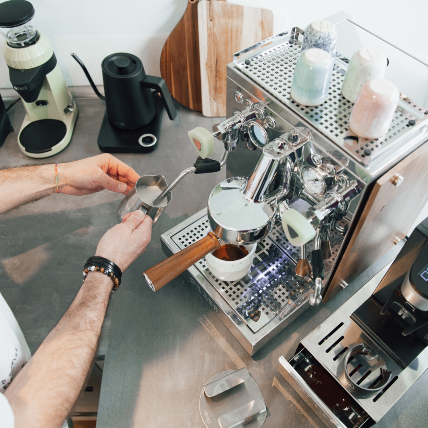 Barista gießt Milch in eine Tasse mit Espresso, Kaffeemaschine im Hintergrund.