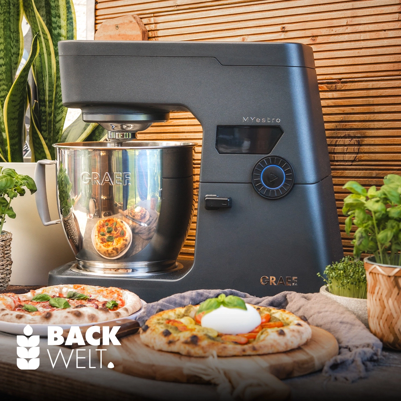 Eine Küchenmaschine von Graef mit Pizzas und Kräutern daneben.