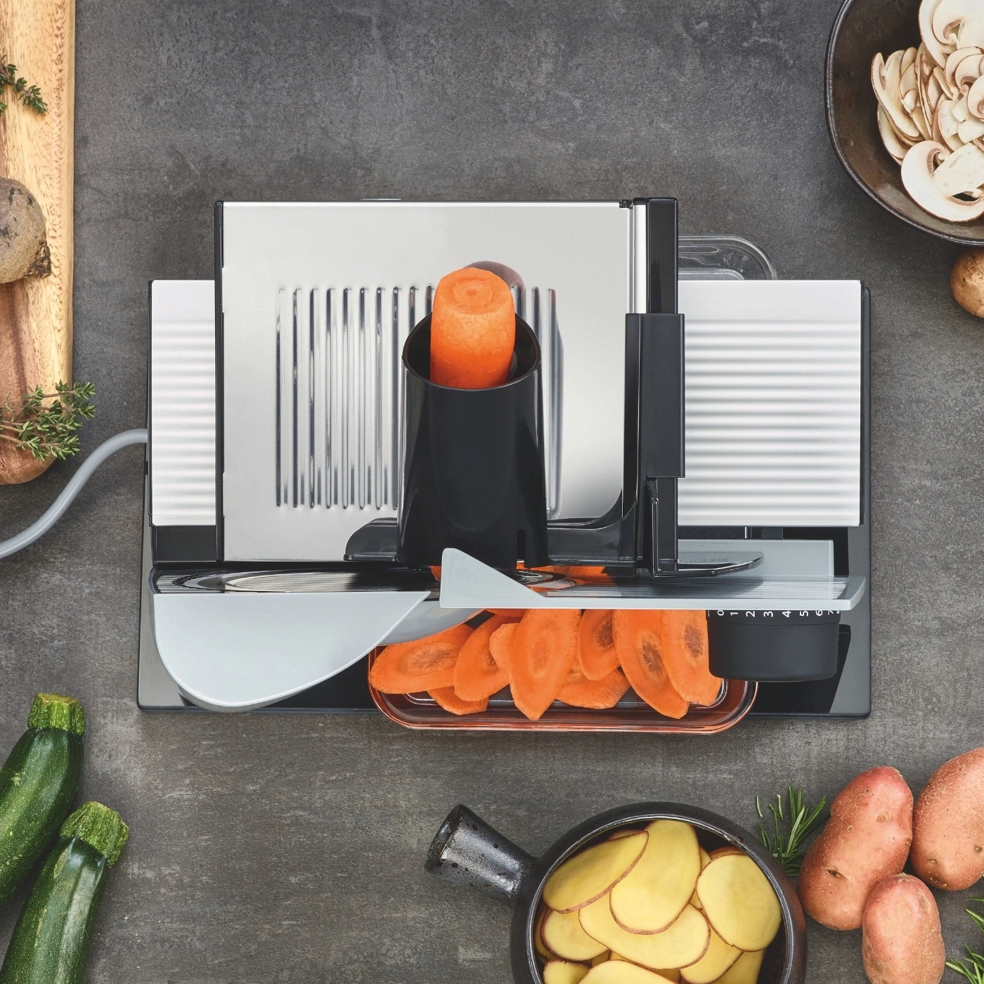 Vegetable slicer with carrots, zucchini, potatoes, and mushrooms on a gray surface.
