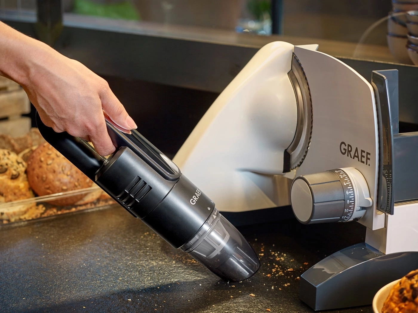 Handheld vacuum cleaner cleaning crumbs near a bread slicer machine.