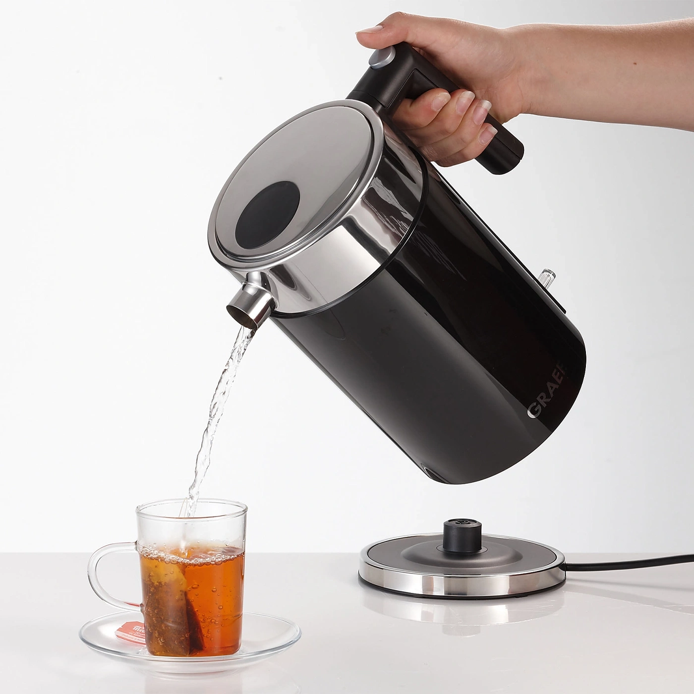 A hand pours water from a black kettle into a teacup with a tea bag.