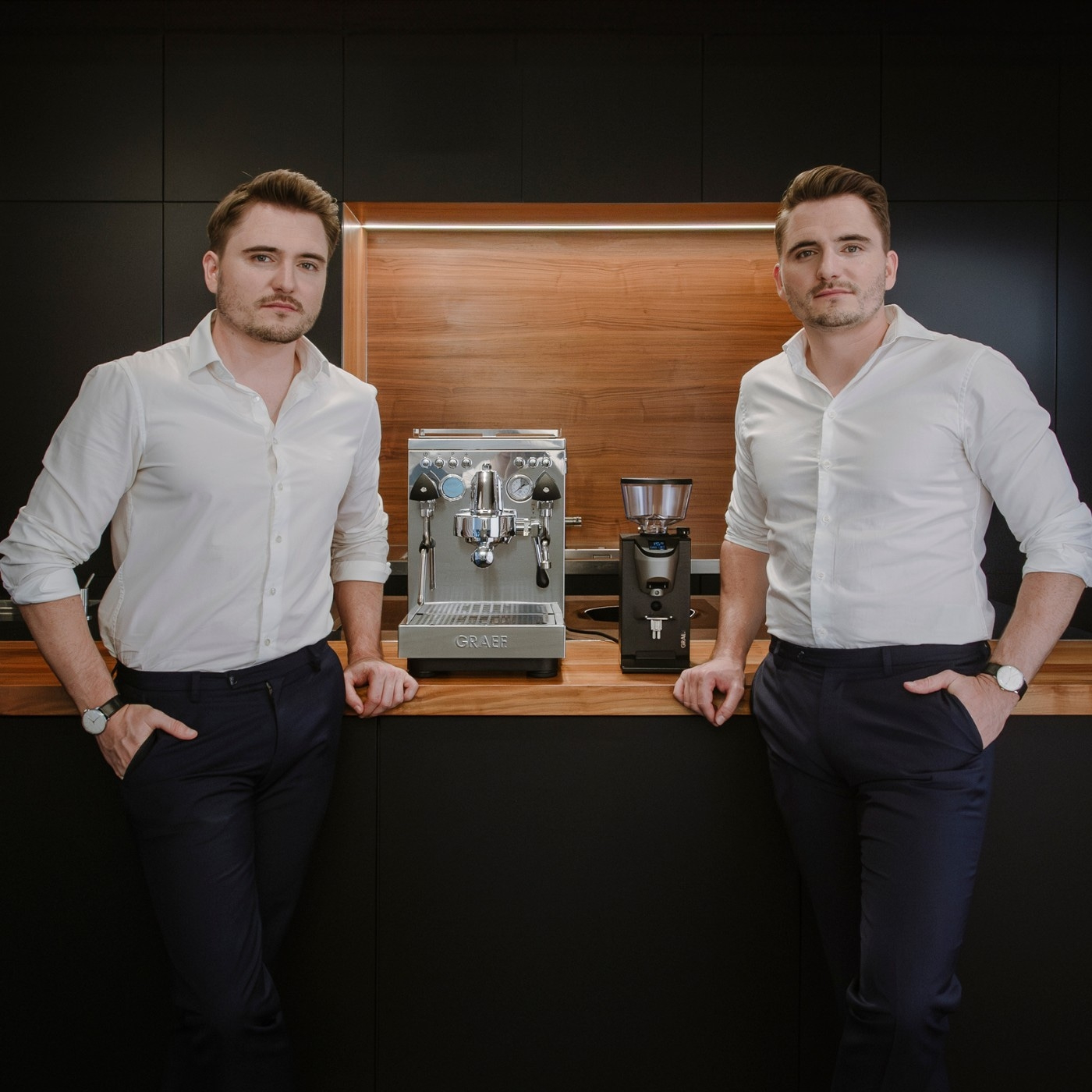 Zwei Männer posieren mit Kaffeemaschine und Mühle in der Küche.