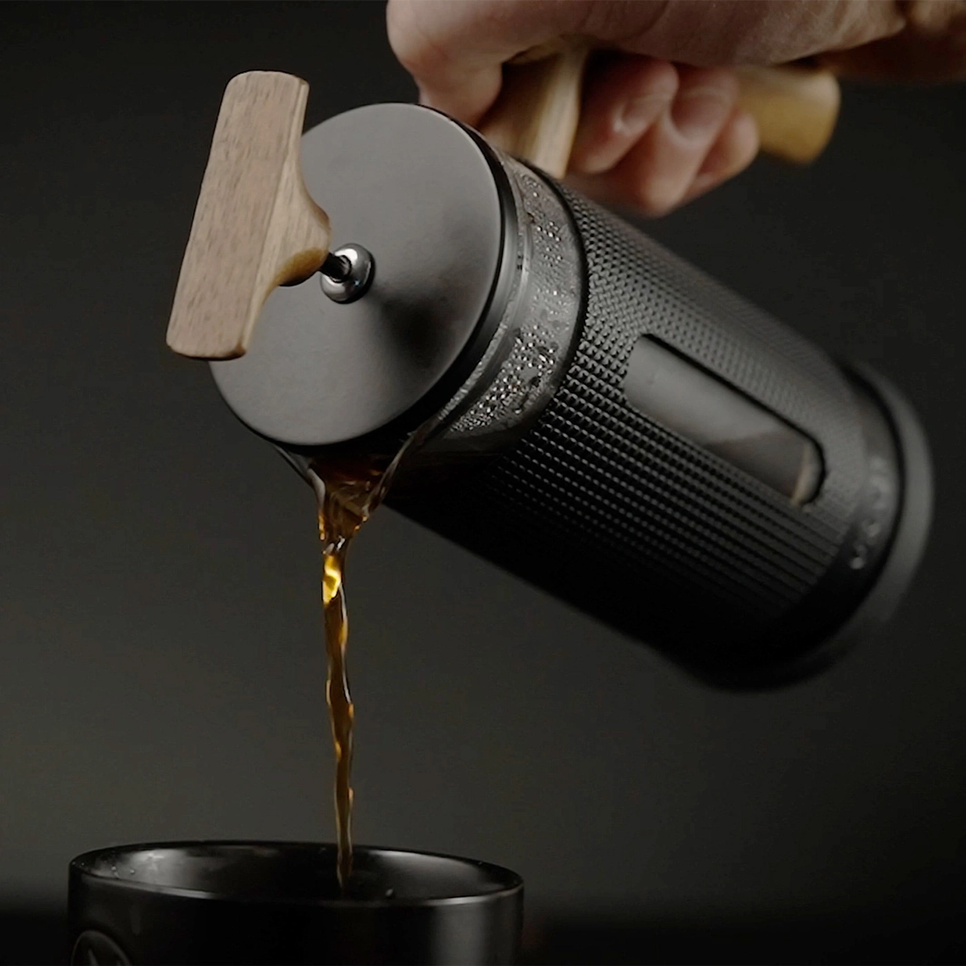 A hand holds a coffee press pouring coffee into a cup.