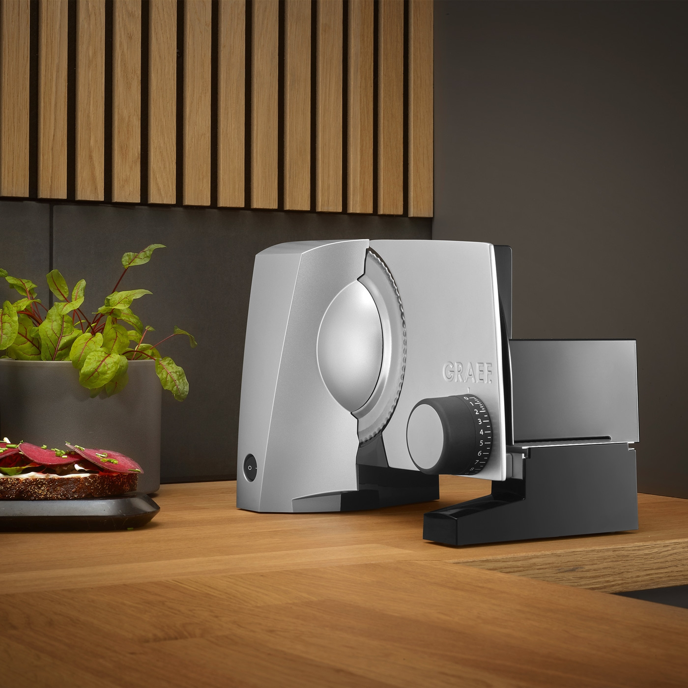 Graef Allesschneider auf Holztisch mit Brot und Pflanze im Hintergrund