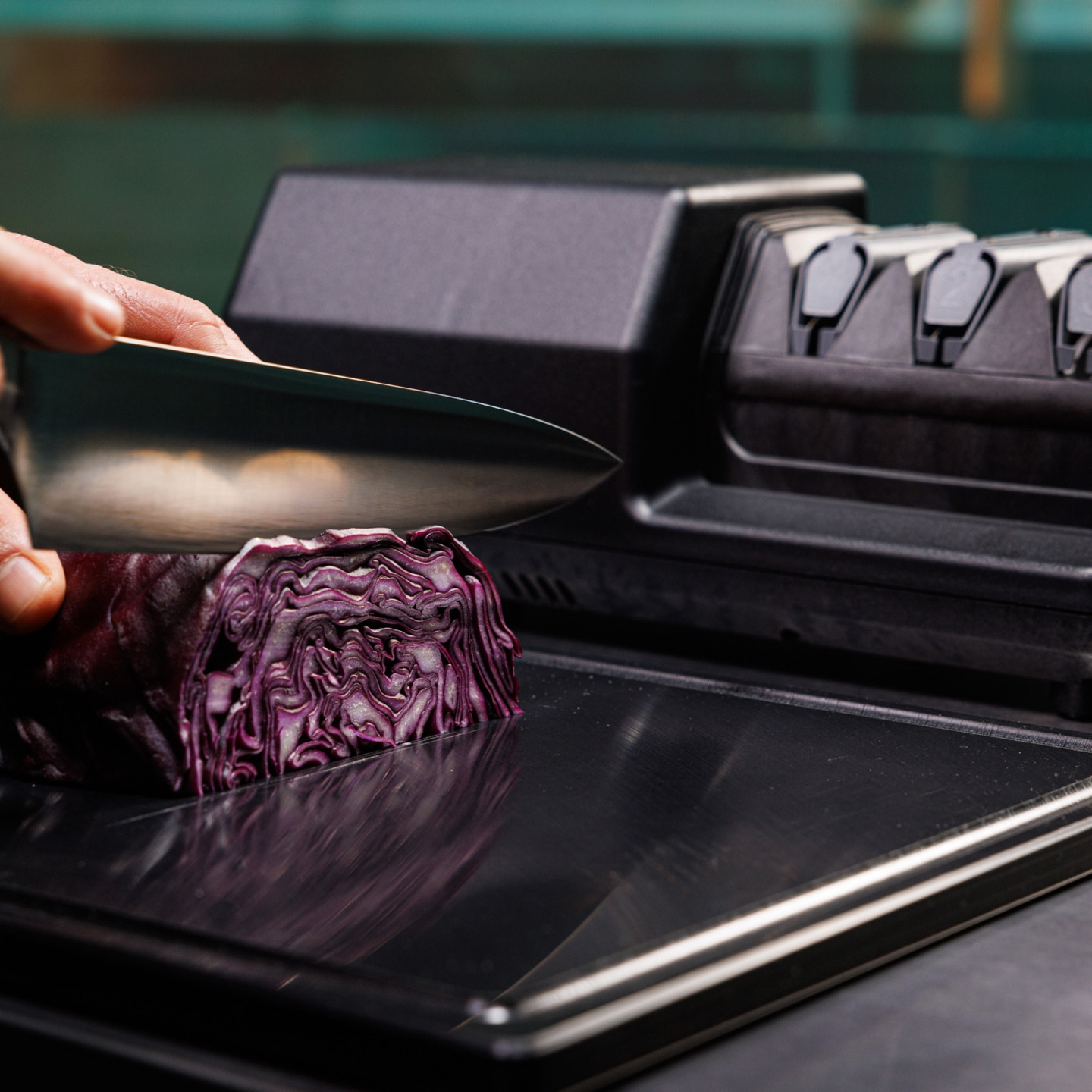 Knife slicing red cabbage on black surface, with a knife sharpener in the background.