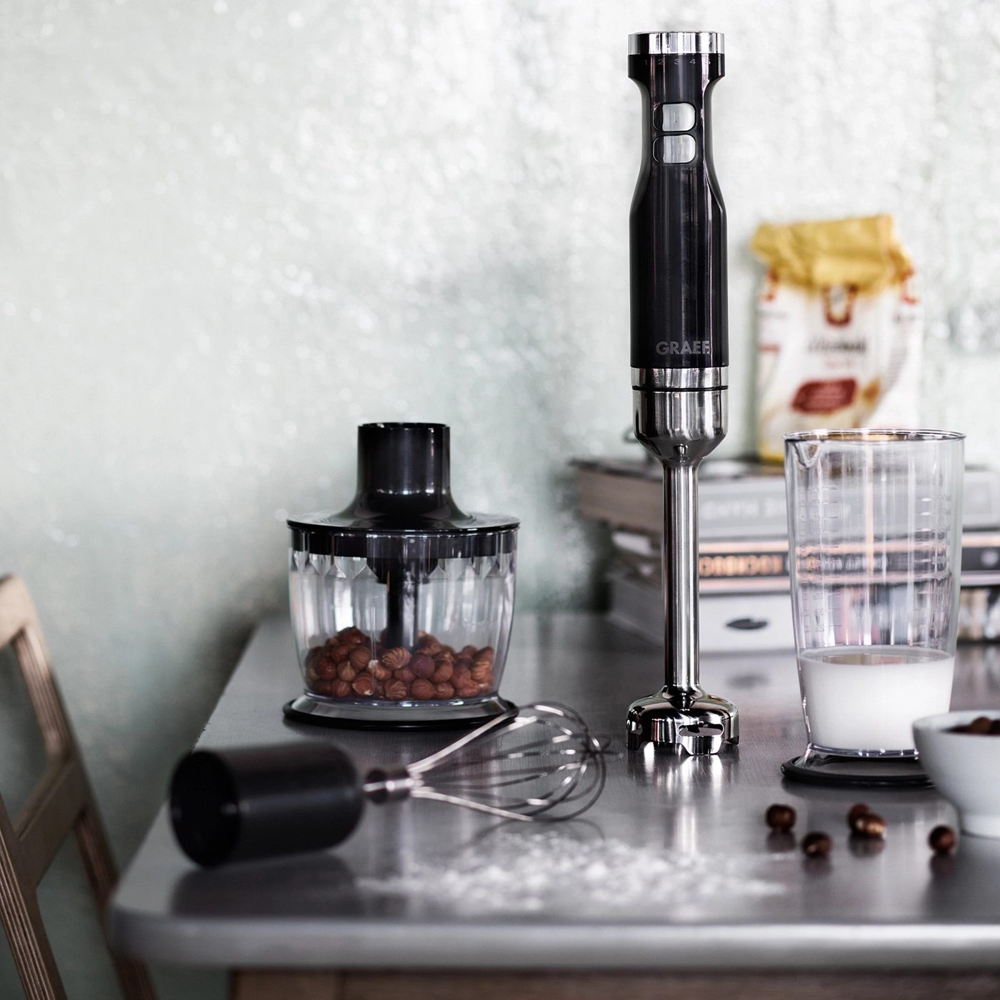 Kitchen utensils on a table: blender, container, milk, nuts, whisk, and ingredients.