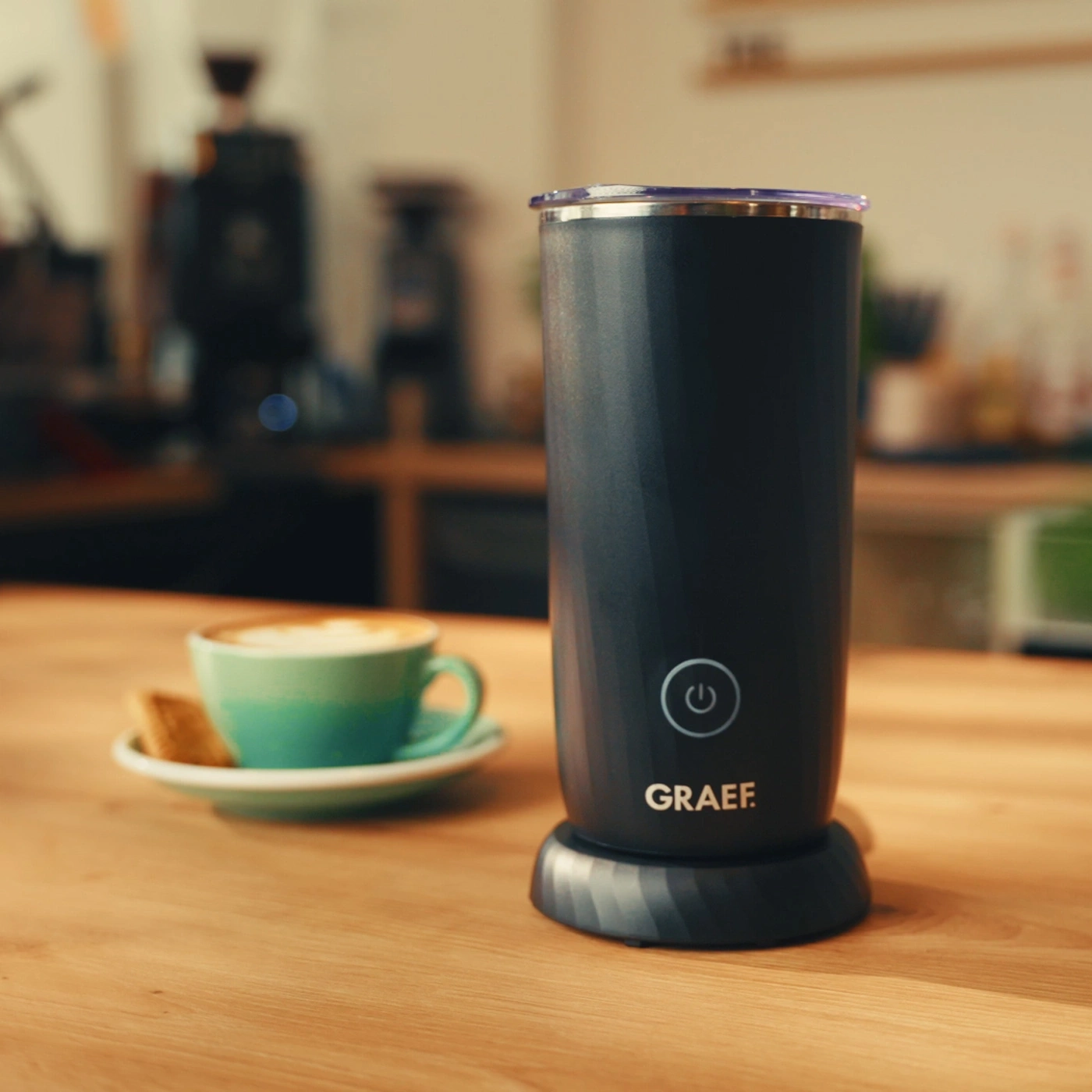 A GRAEF milk frother next to a cup of coffee on a wooden table.