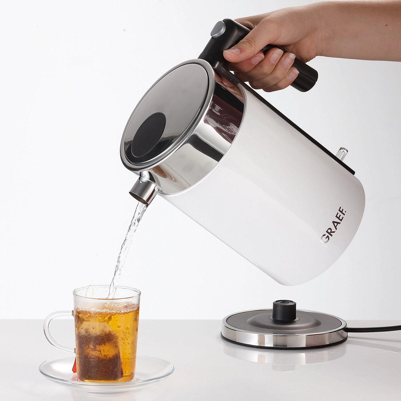 White kettle pouring hot water into a teacup with a tea bag inside.