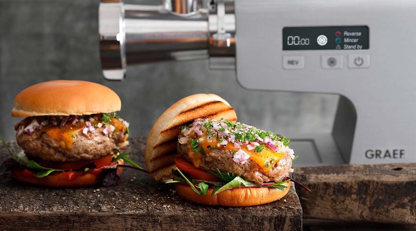 Burger mit Käse und Zwiebeln neben Fleischwolf, auf Holzbrett