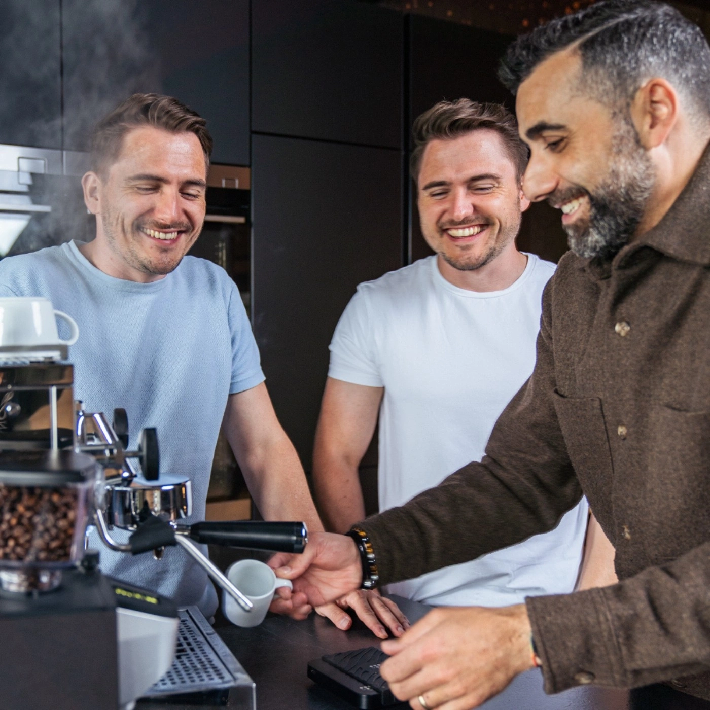 Drei Männer machen Kaffee zusammen, lächeln und haben Spaß in der Küche.