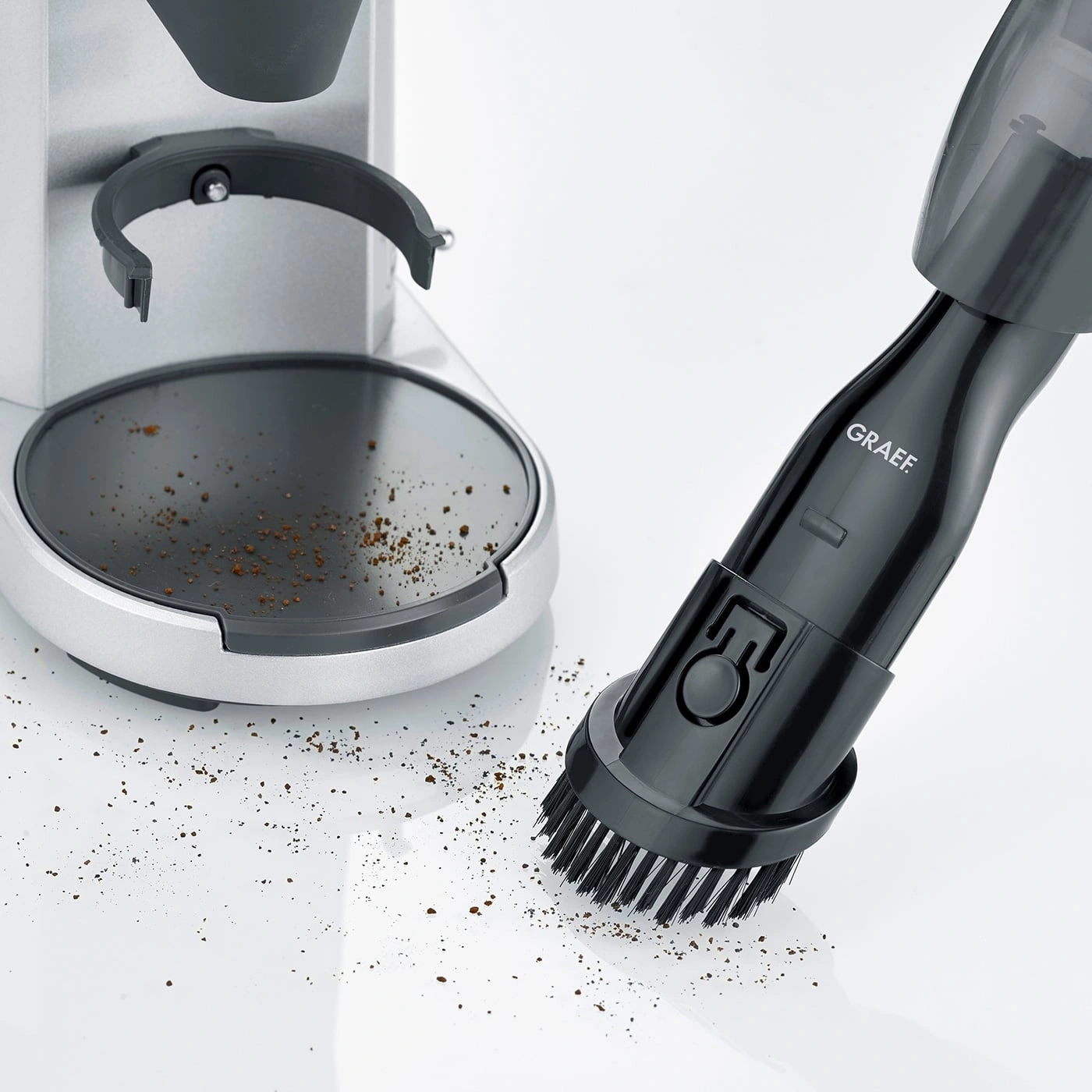 Coffee grinder and vacuum cleaner with brush cleaning coffee grounds on white surface.