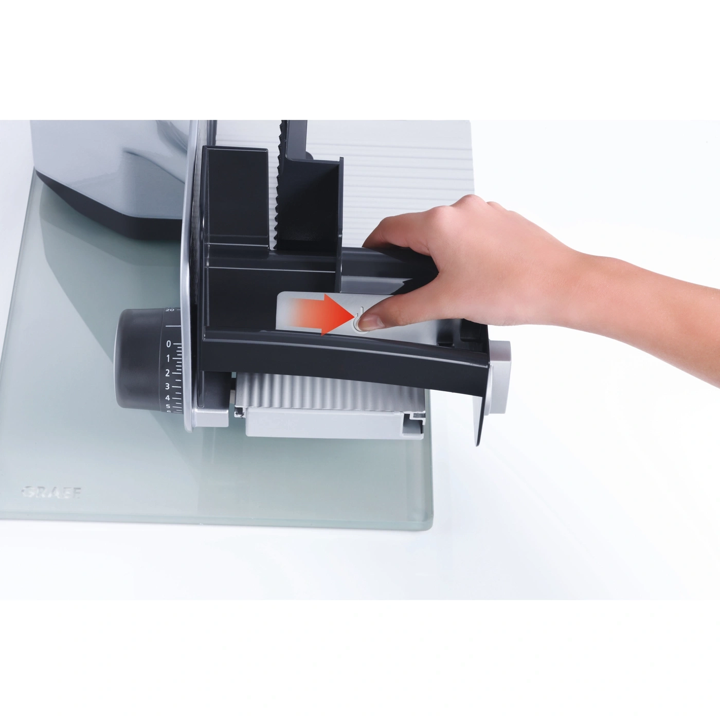 Close-up of a meat slicer with a hand pressing a button to operate it, with an arrow.