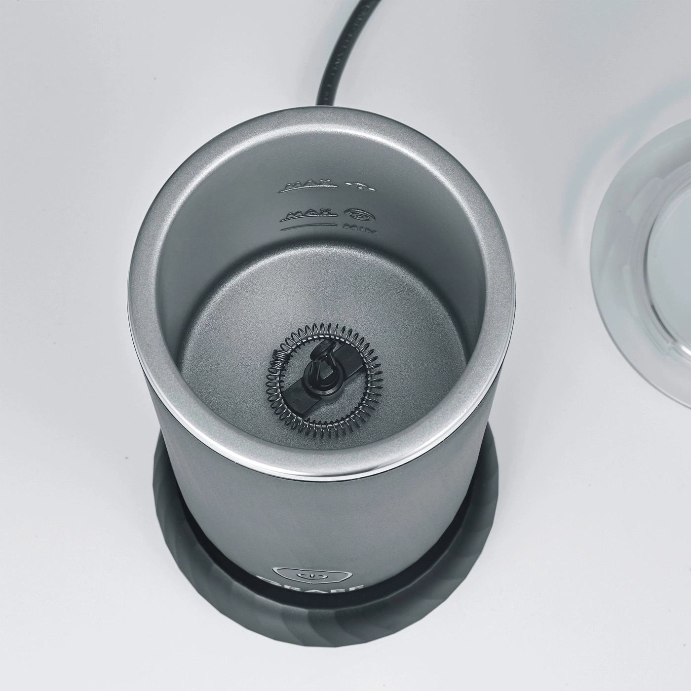 Grey milk frother with whisk and markings on a white surface, top view