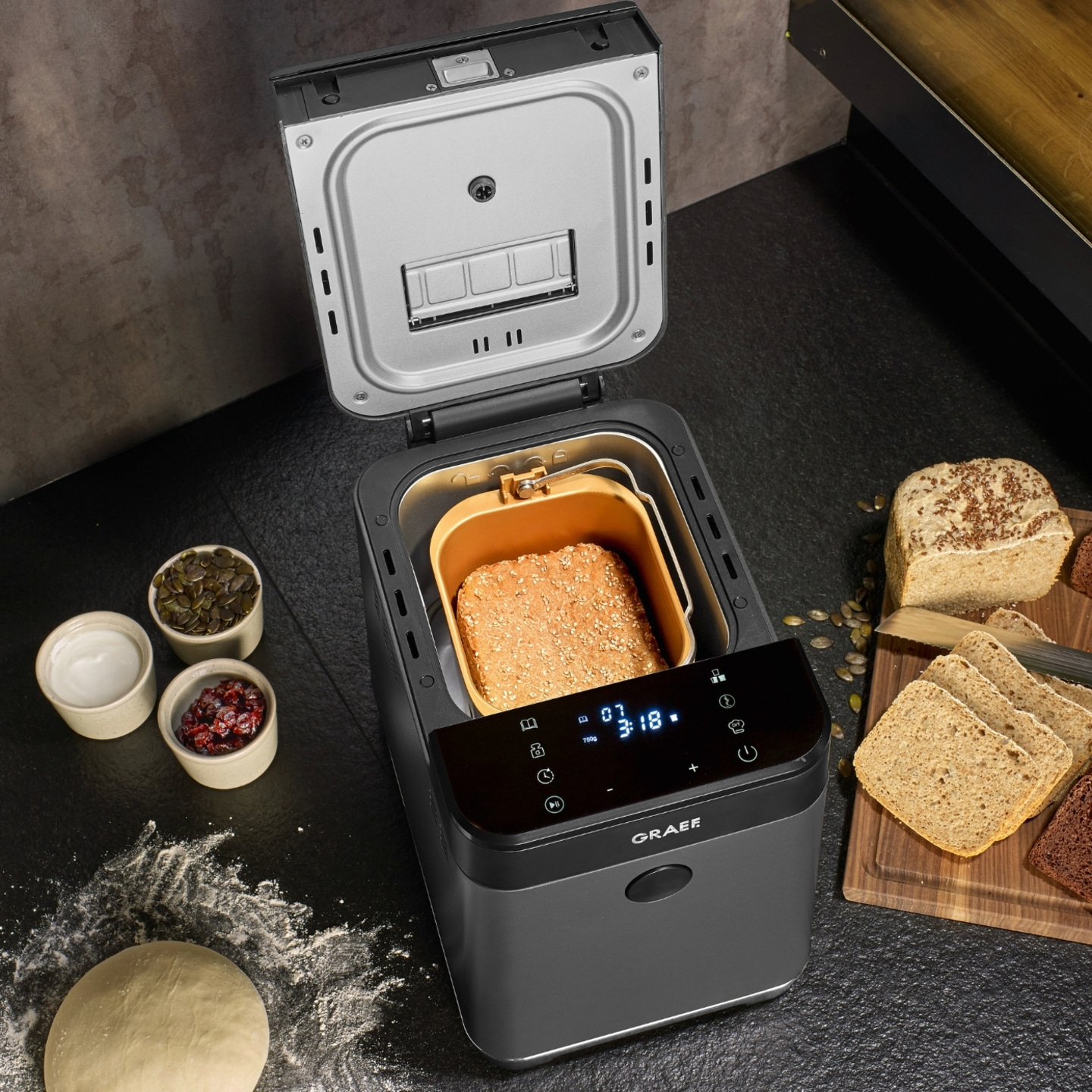 Bread maker with bread, ingredients, and loaf on wooden board
