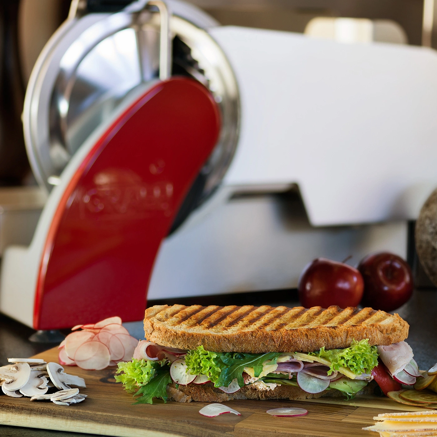 Sandwich mit Gemüse und Fleisch, neben einer Aufschnittmaschine und Äpfeln