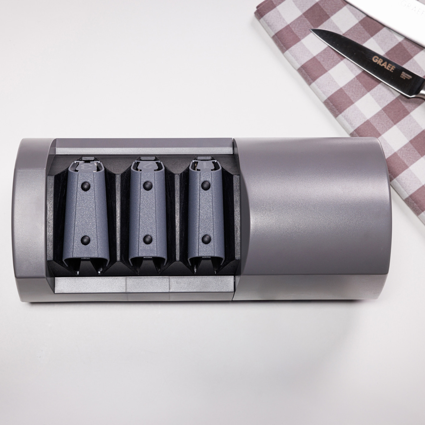 Graues Messer-Schärfgerät mit drei Schlitzen und einem Messer auf einem karierten Tuch.