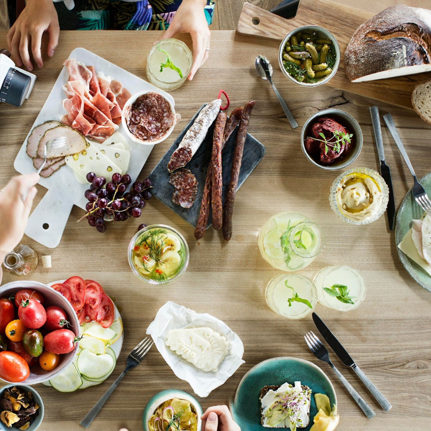 Holztisch mit Essen: Wurst, Käse, Gemüse, Getränke, Brot, Menschen.