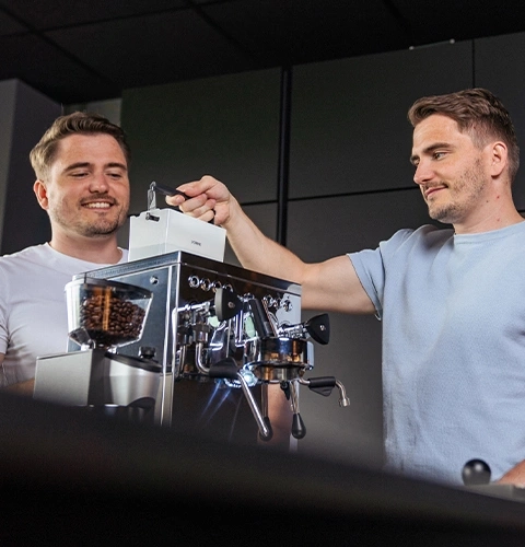 Two men at an espresso machine, preparing coffee and smiling together.