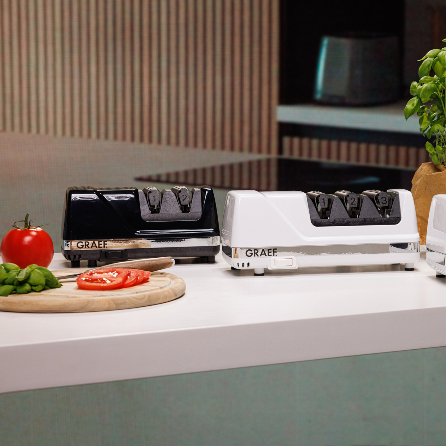 Two electric knife sharpeners, one black, one white, on a countertop with tomatoes and basil.