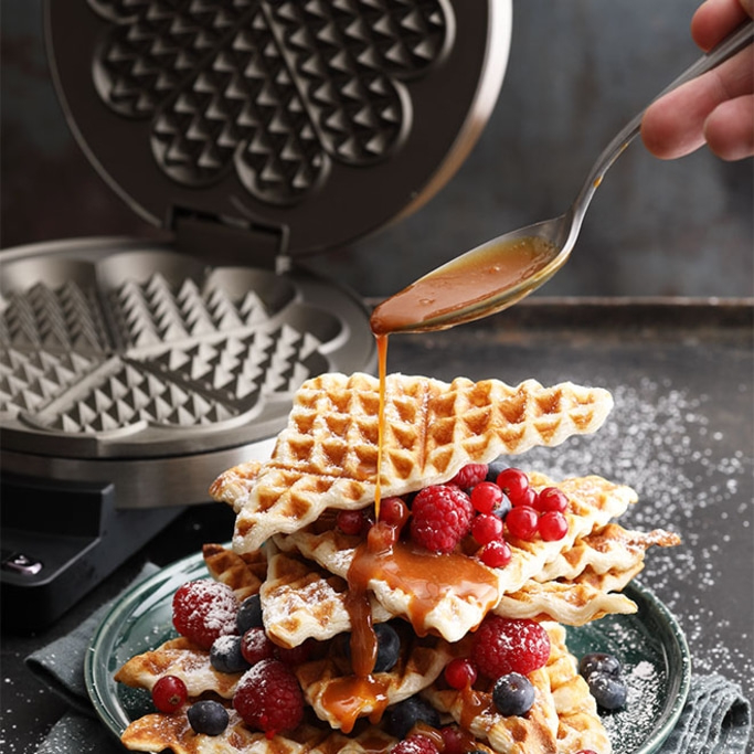 Waffeln mit Beeren und Sirup, die von einer Hand mit einem Löffel übergossen werden.
