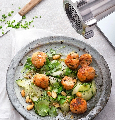 Fleischbällchen-Salat mit Erdnüssen und Gurken auf einem Teller mit Messer und Fleischwolf