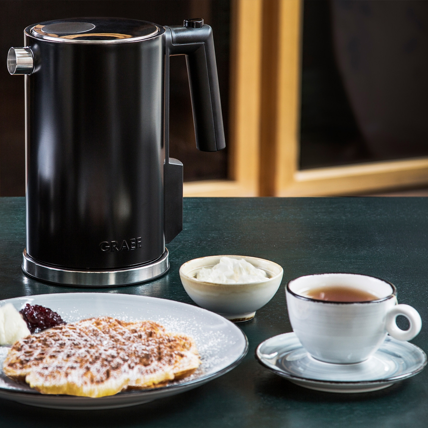 Schwarzer Wasserkocher, Waffel, Tee, Sahne und Marmelade auf dem Tisch.