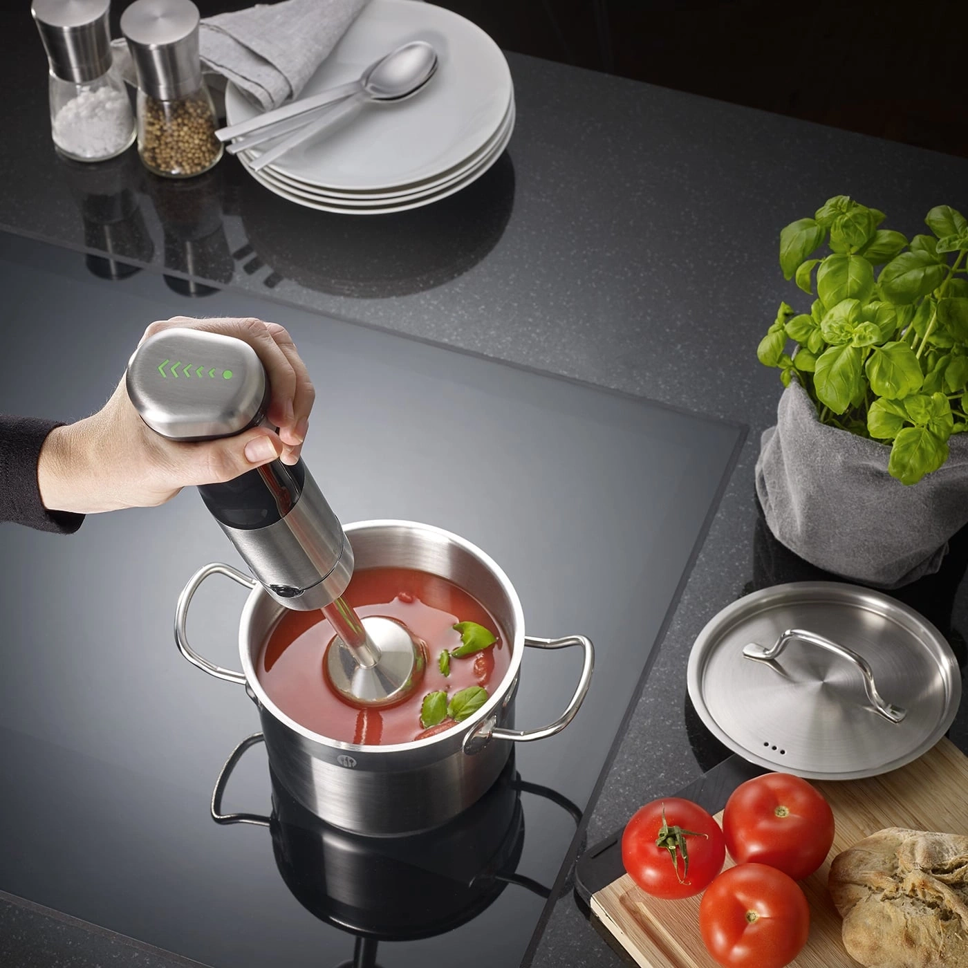 Hand blender in a pot of tomato soup, tomatoes and basil next to it.