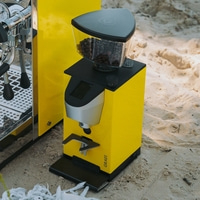 A yellow coffee grinder and espresso machine stand on the beach in the sand.