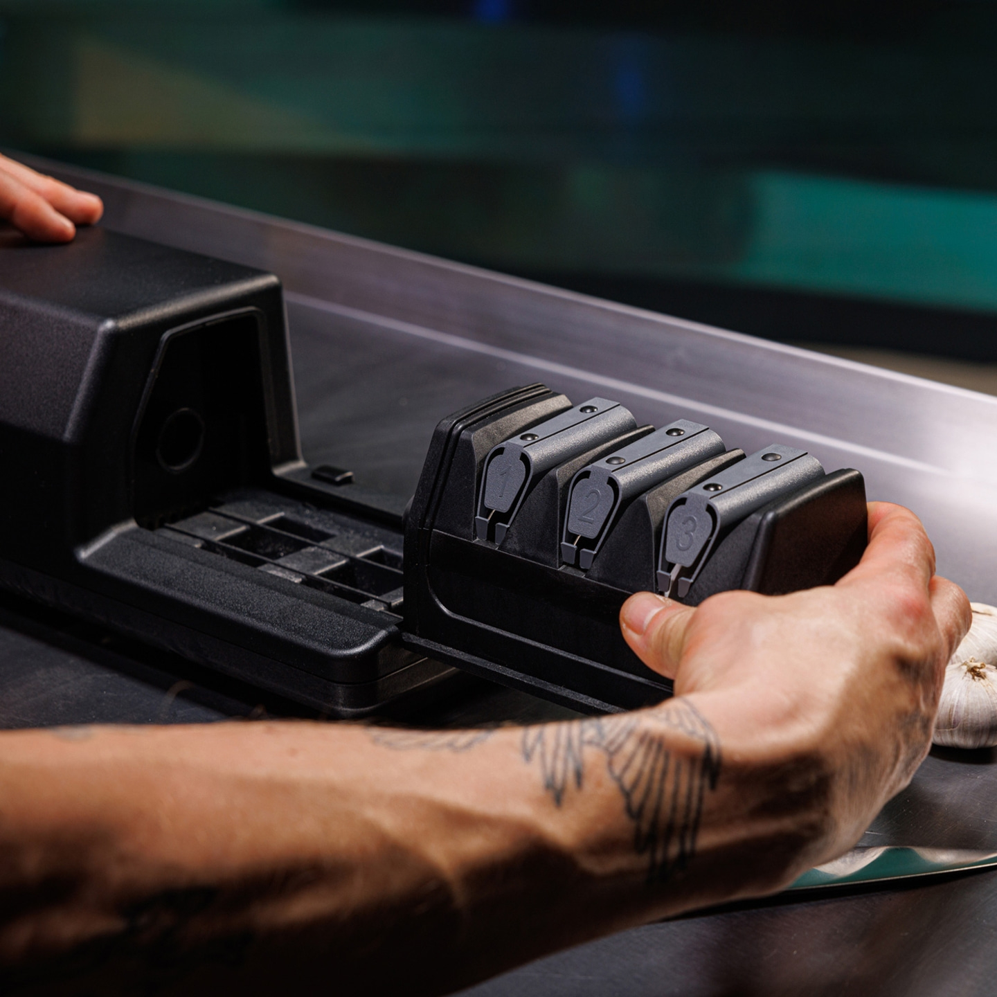 Person holding a black knife sharpener on a metal surface with tattoos.