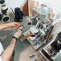 Barista gießt Milch in eine Tasse mit Espresso, Kaffeemaschine im Hintergrund.