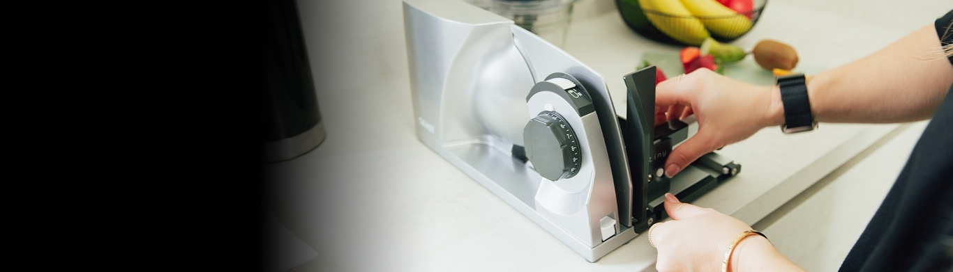 Person operating a slicer machine in kitchen with fruits and vegetables