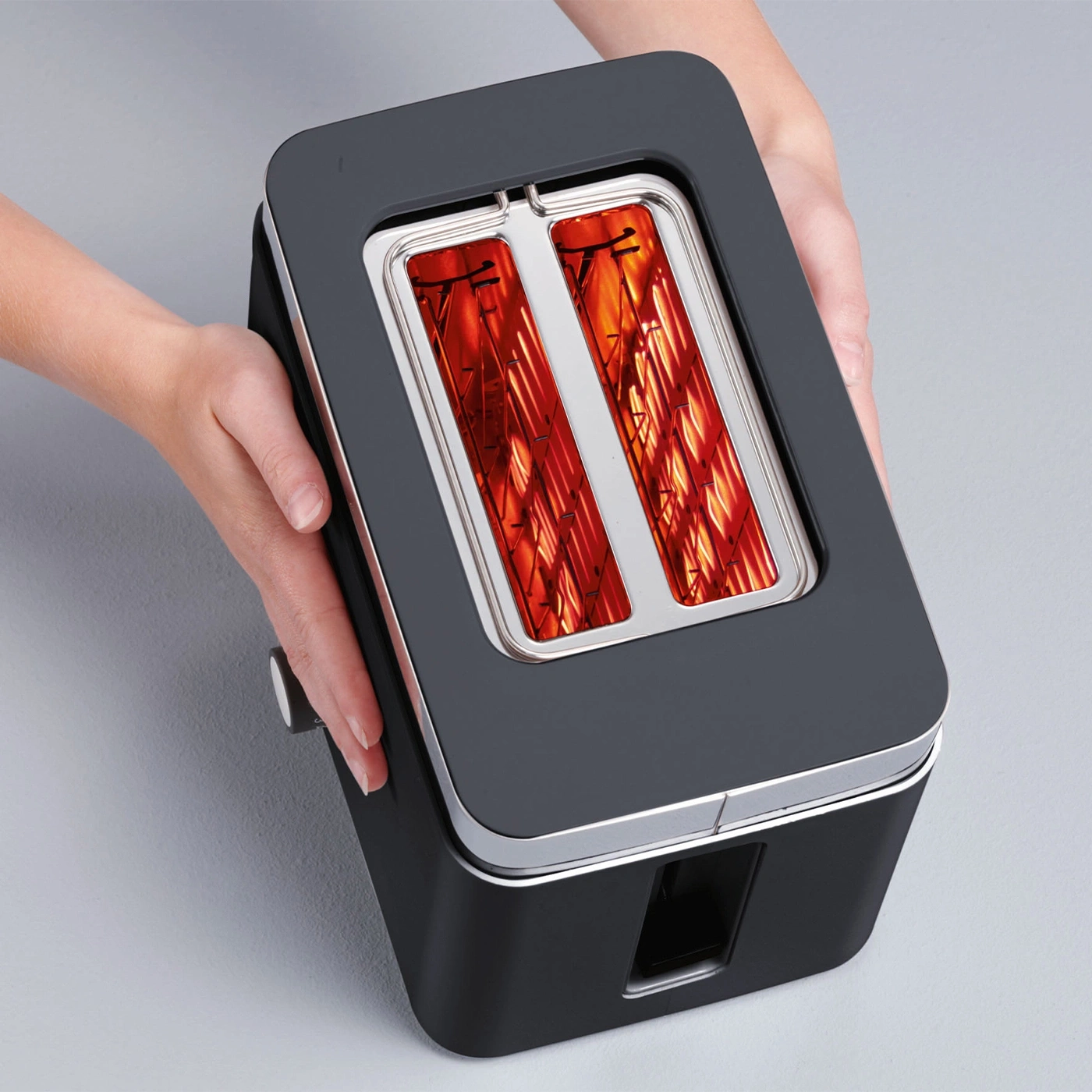 Toaster in Händen, schwarz, mit silbernen Akzenten, Toastschlitze leuchten rot.