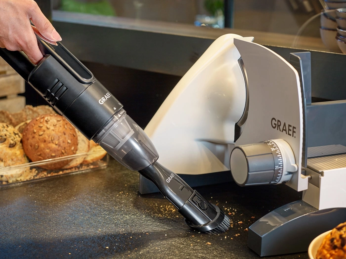 Handheld vacuum cleaner cleaning bread crumbs next to a Graef slicer.